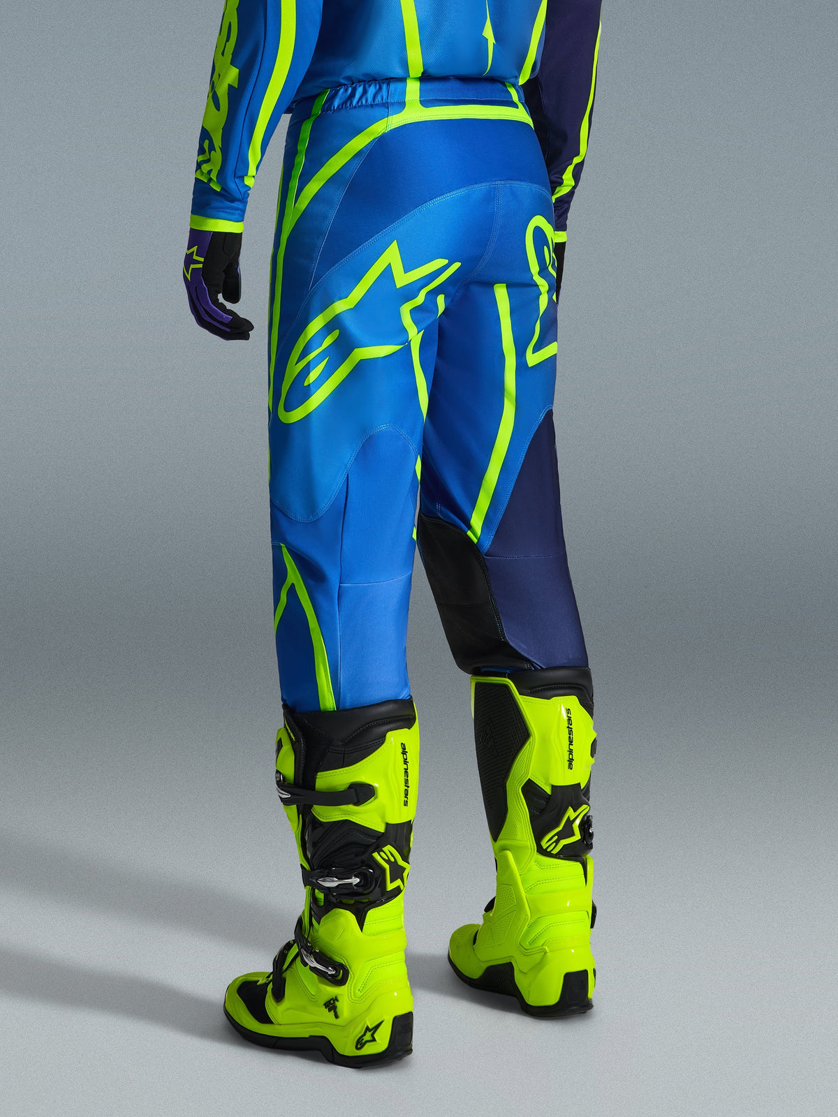 Alpinestars 2026 Fluid Apex Pants, pantalones de motocross, azul y amarillo flúor, vista trasera del piloto con equipamiento a juego y botas amarillo flúor, con logotipos llamativos de Alpinestars y diseño ergonómico