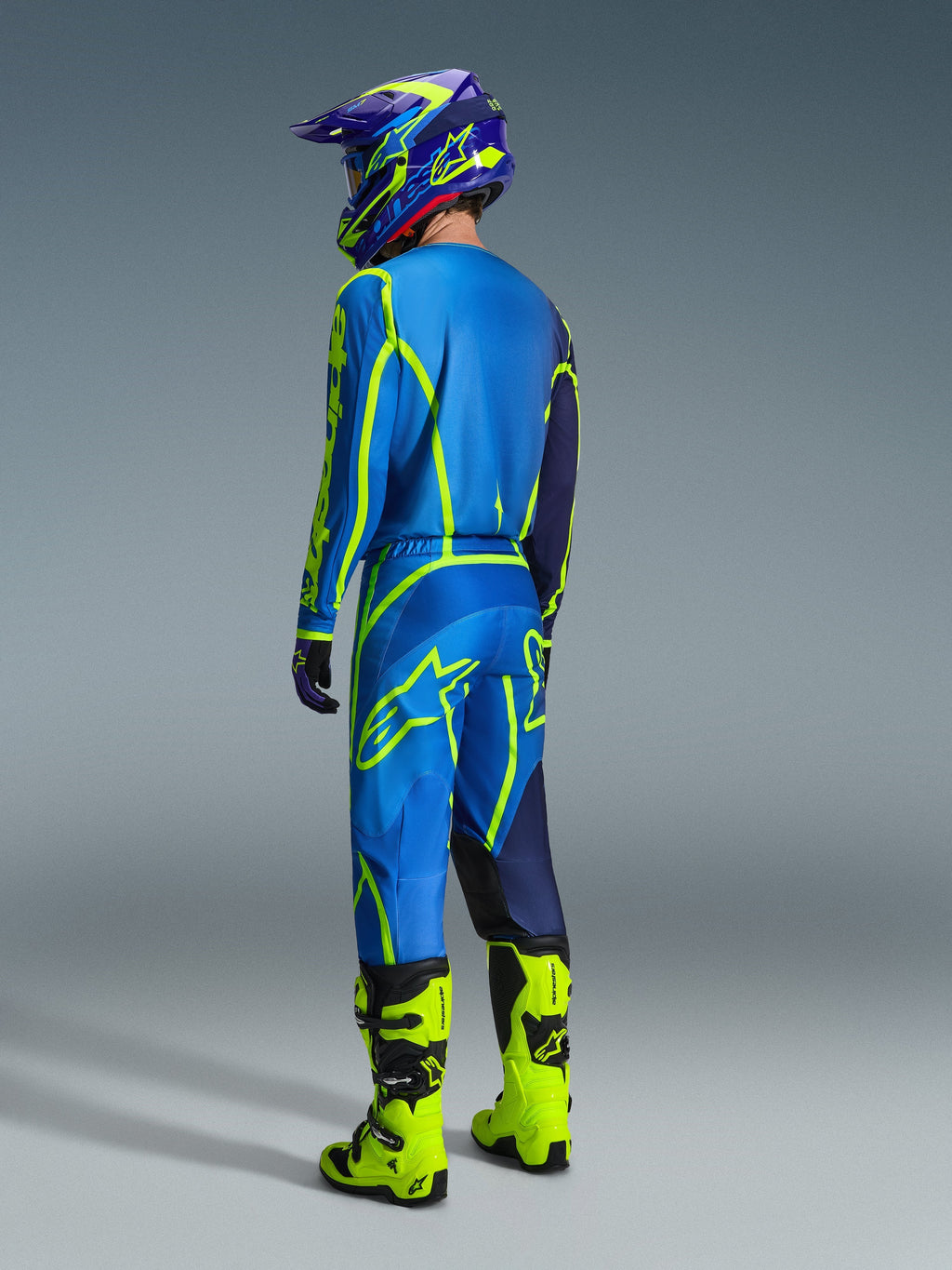 Piloto de motocross visto desde atrás con pantalones Alpinestars Fluid Apex 2026 en azul y amarillo flúor, jersey y casco a juego en azul y amarillo, y botas amarillo neón brillante, equipo de competición coordinado para rendimiento off-road