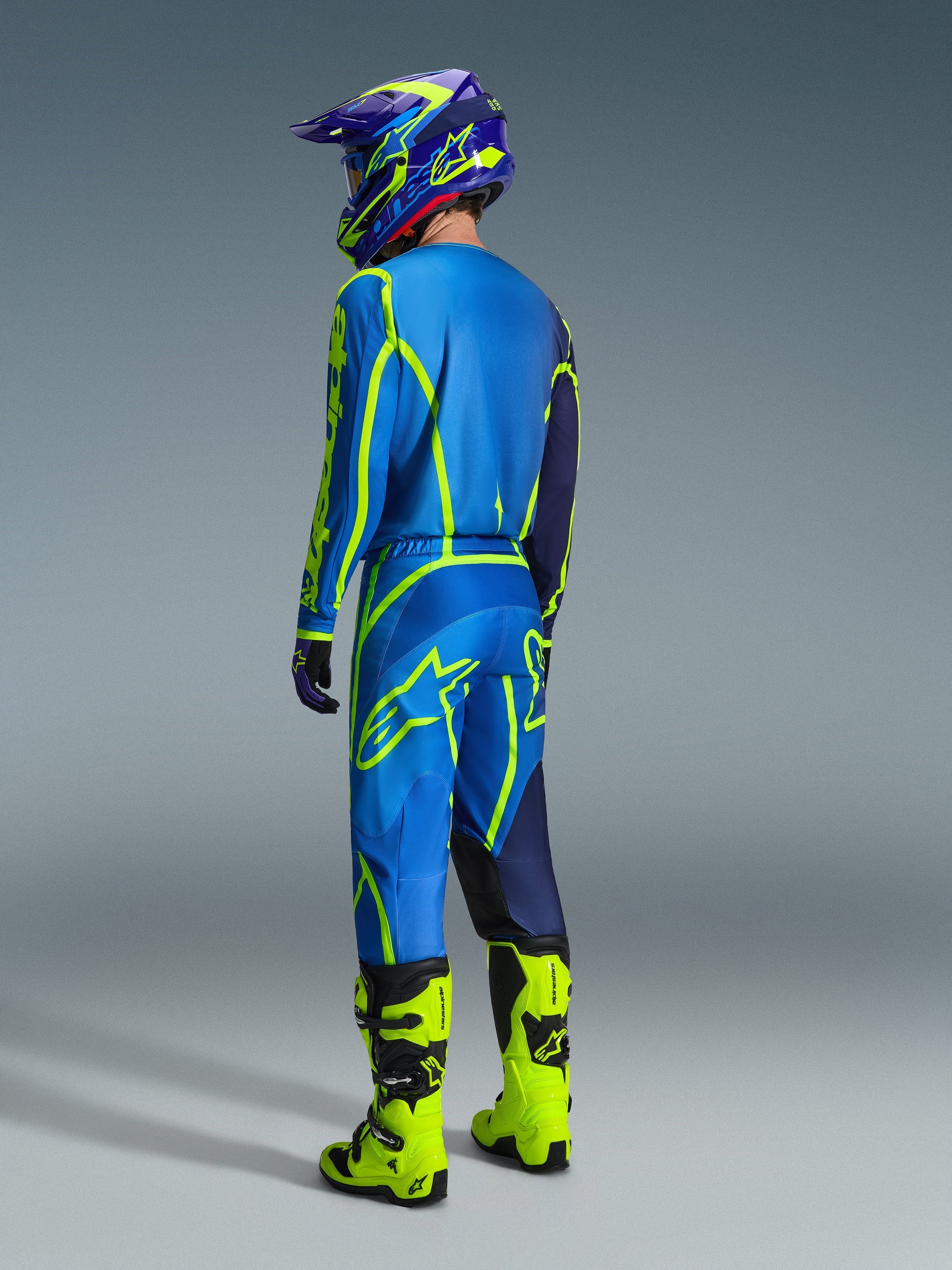 Piloto de motocross visto desde atrás con pantalones Alpinestars Fluid Apex 2026 en azul y amarillo flúor, jersey y casco a juego en azul y amarillo, y botas amarillo neón brillante, equipo de competición coordinado para rendimiento off-road