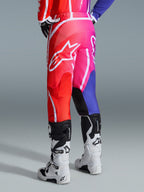 Pantalones Alpinestars 2026 Fluid Apex, pantalones de motocross, multicolor rojo, rosa y púrpura, vista trasera de piloto con botas de motocross blancas y negras, con logo grande de Alpinestars en blanco y diseño de degradado vibrante