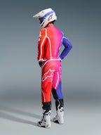 Piloto de motocross con equipamiento Alpinestars 2026 Fluid Apex, jersey y pantalones multicolores con un degradado en rojo, rosa y morado y logotipos blancos, casco blanco y botas blancas, conjunto coordinado para carreras todoterreno