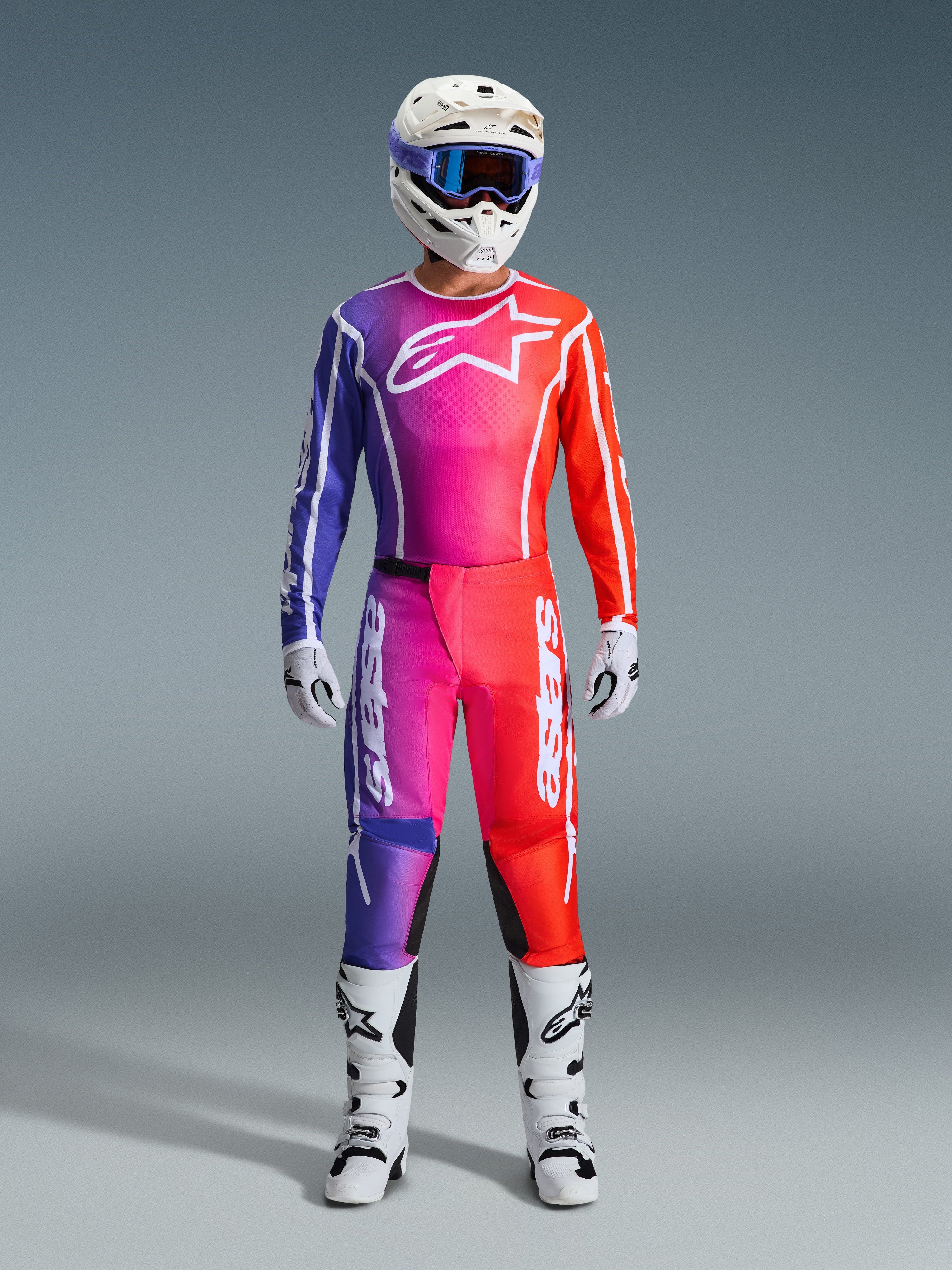 Piloto de motocross con pantalones Fluid Apex 2026 y jersey a juego, degradado de púrpura y naranja, casco blanco con gafas, botas y guantes blancos, equipamiento de carreras Alpinestars coordinado para rendimiento off-road.