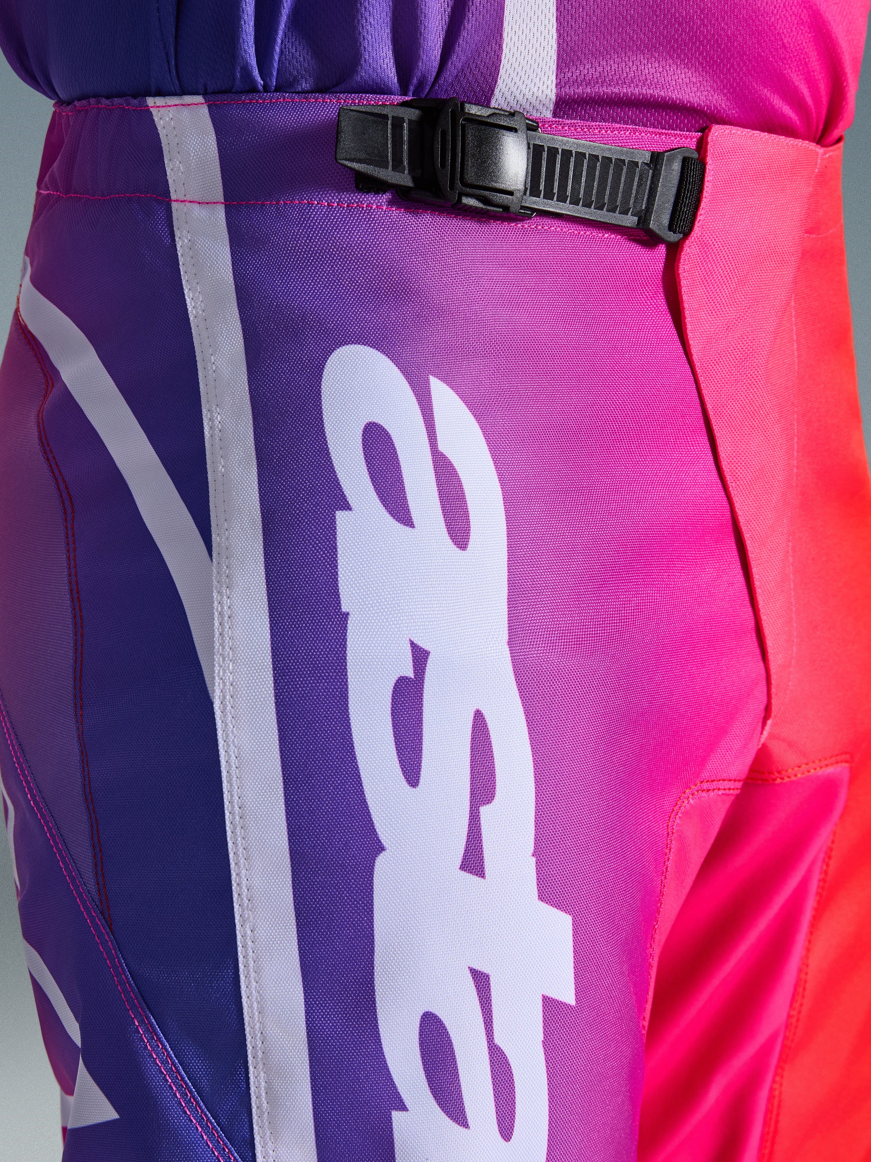 Alpinestars 2026 Fluid Apex Pants, pantalones de motocross, multicolor púrpura, rosa y rojo, primer plano de la zona de la cintura y la cadera con sistema de cierre de trinquete negro y gran logotipo vertical de Alpinestars en blanco, diseño con degradado vibrante y textura de polifabric duradera