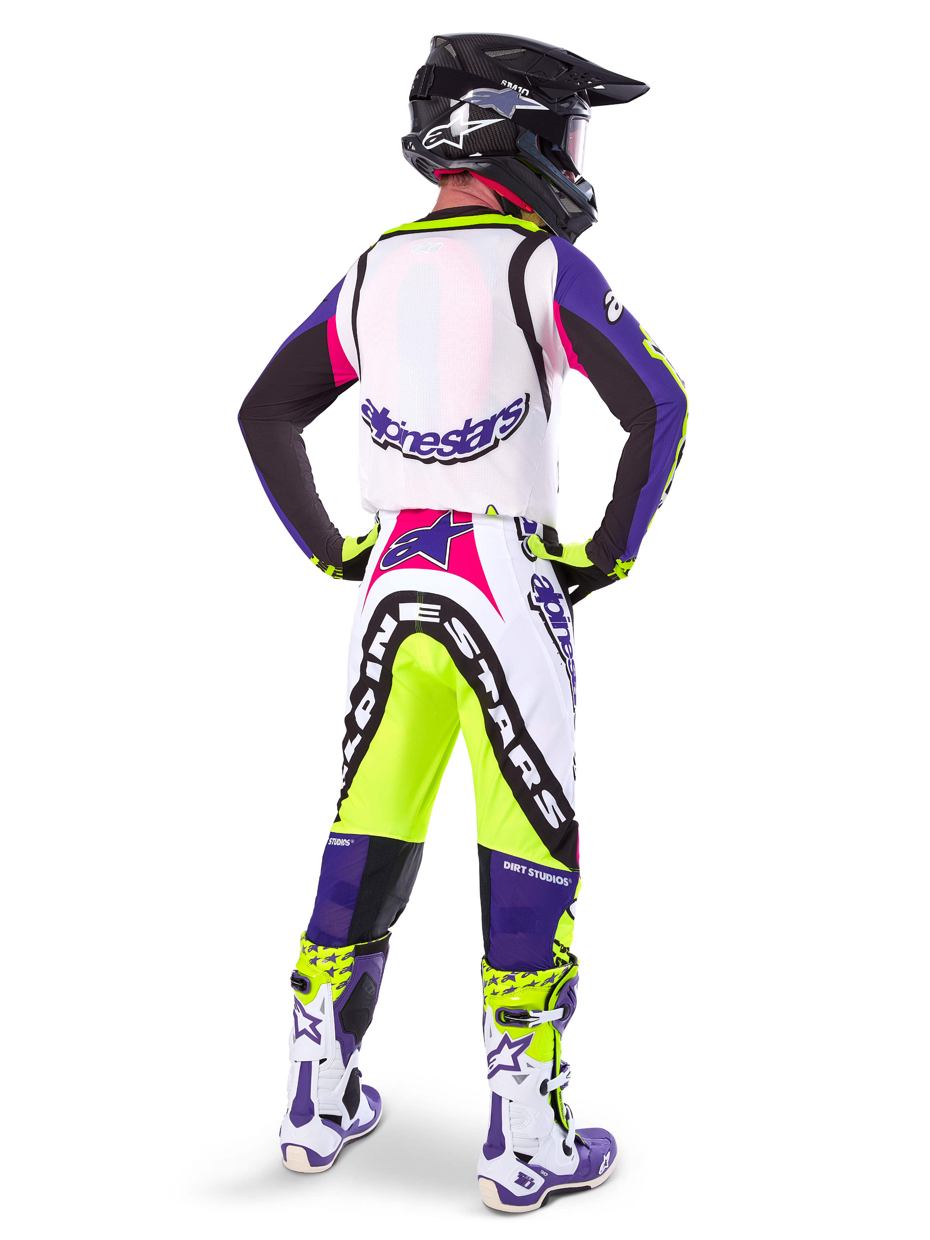 Motocross rider wearing Alpinestars Limited Edition Supertech Dirt Studios® Pants in White Purple and Yellow Fluo, seen from the back with matching jersey, boots, and black carbon helmet, professional off-road racing gear with bold graphics and neon accents.