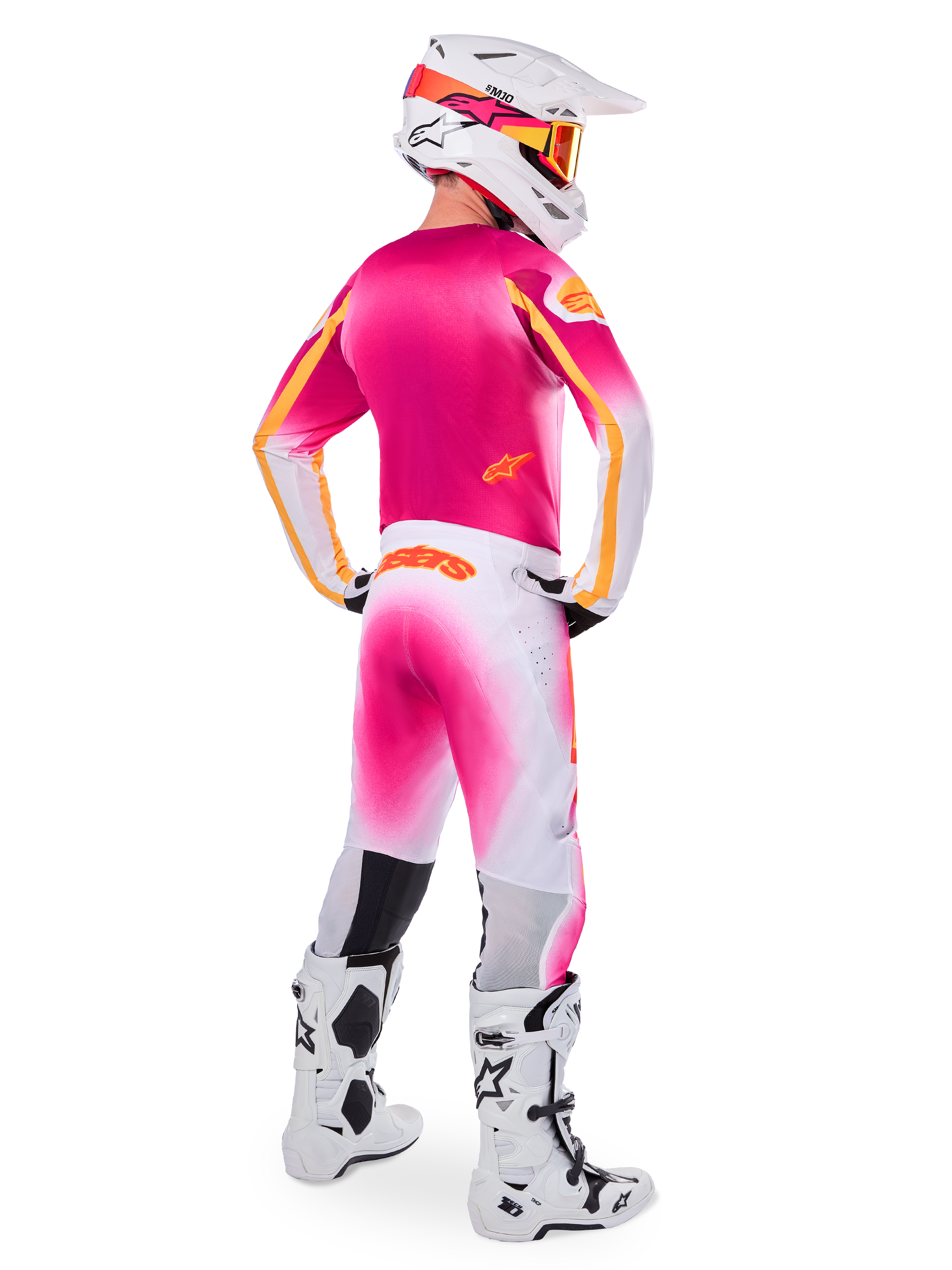 Piloto de motocross visto desde atrás con el conjunto de equipamiento de edición limitada Supertech AFD MIG, jersey rosa con detalles naranjas, pantalones blancos con degradado rosa y marca Alpinestars naranja, casco blanco con gráficos rosas y gafas naranjas, botas blancas y negras, kit de competición profesional para todoterreno