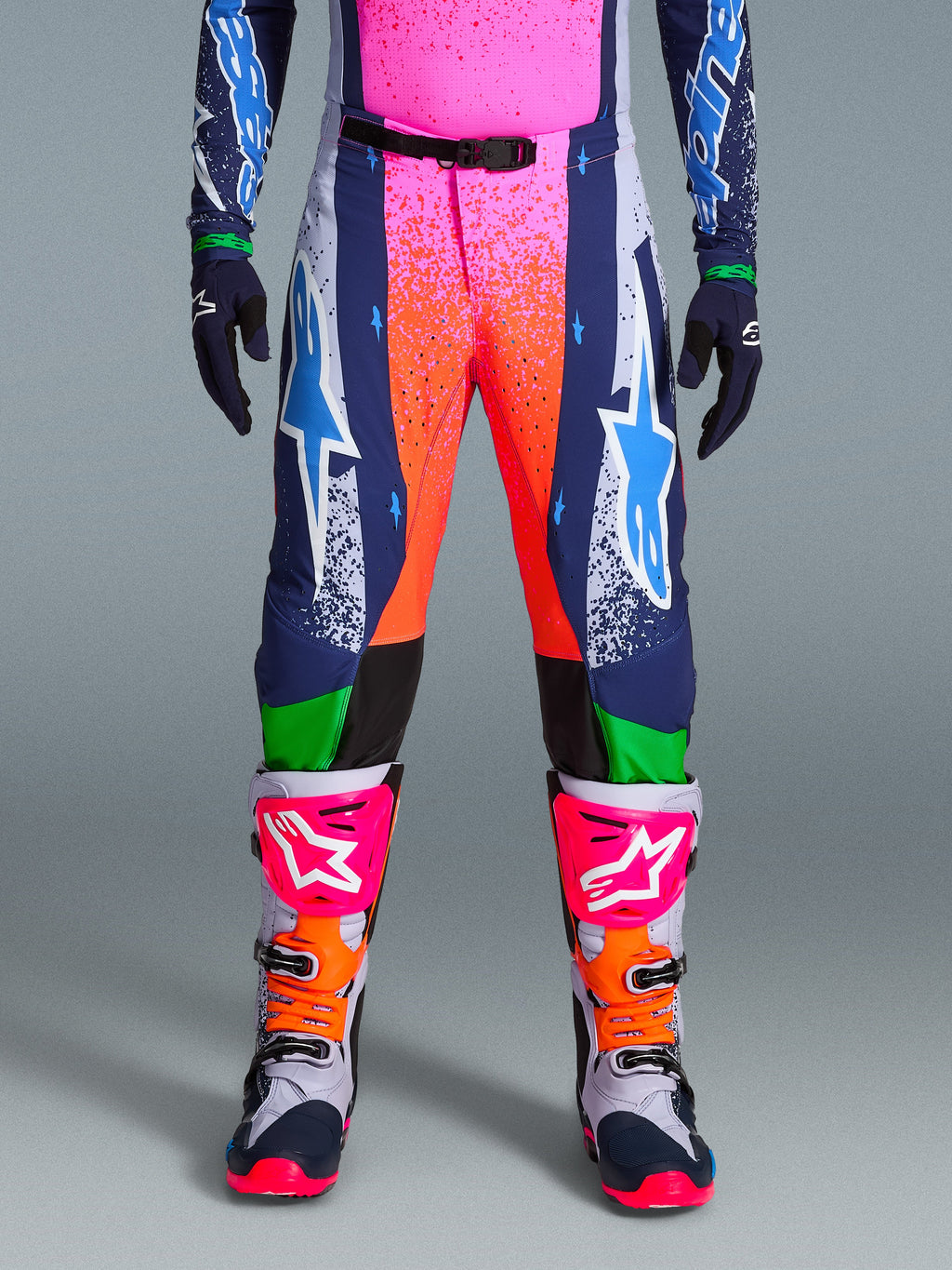Alpinestars Limited Edition Supertech Records Pants, pantalones de motocross, multicolor con vibrantes gráficos de salpicaduras en rosa y naranja, paneles azul marino con logotipos de estrellas azules y detalles verdes, usados con jersey a juego y botas de motocross grises, rosas y naranjas