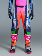 Alpinestars Limited Edition Supertech Records Pants, pantalones de motocross, multicolor con vibrantes gráficos de salpicaduras en rosa y naranja, paneles azul marino con logotipos de estrellas azules y detalles verdes, usados con jersey a juego y botas de motocross grises, rosas y naranjas