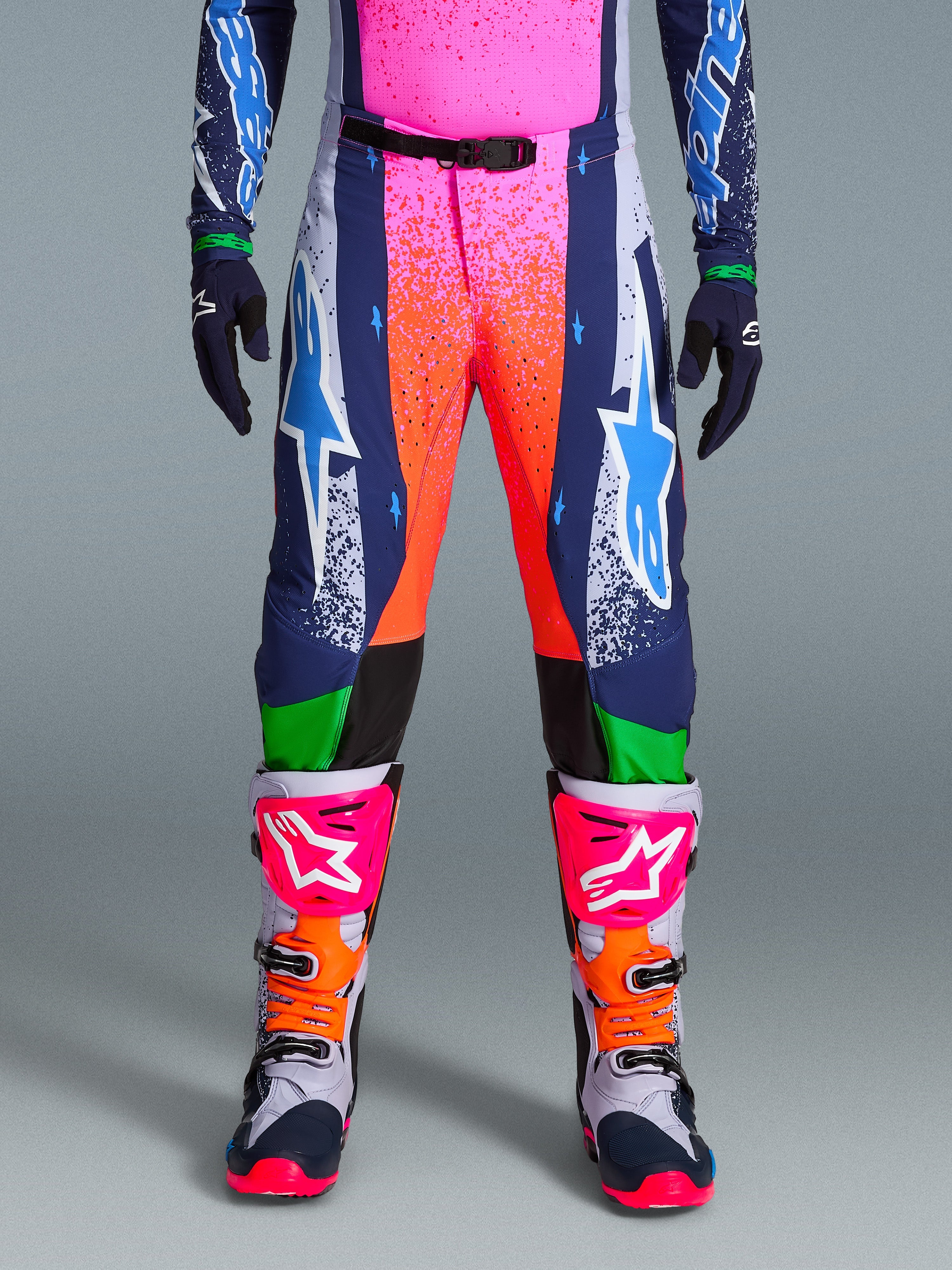 Alpinestars Limited Edition Supertech Records Pants, pantalones de motocross, multicolor con vibrantes gráficos de salpicaduras en rosa y naranja, paneles azul marino con logotipos de estrellas azules y detalles verdes, usados con jersey a juego y botas de motocross grises, rosas y naranjas