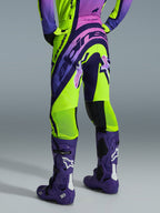 Pantalones Alpinestars Supertech Vista 2026, pantalones de motocross, amarillo, púrpura y negro, vista trasera combinados con jersey a juego y botas de motocross moradas, con logotipos llamativos y zonas de malla transpirable en las pantorrillas
