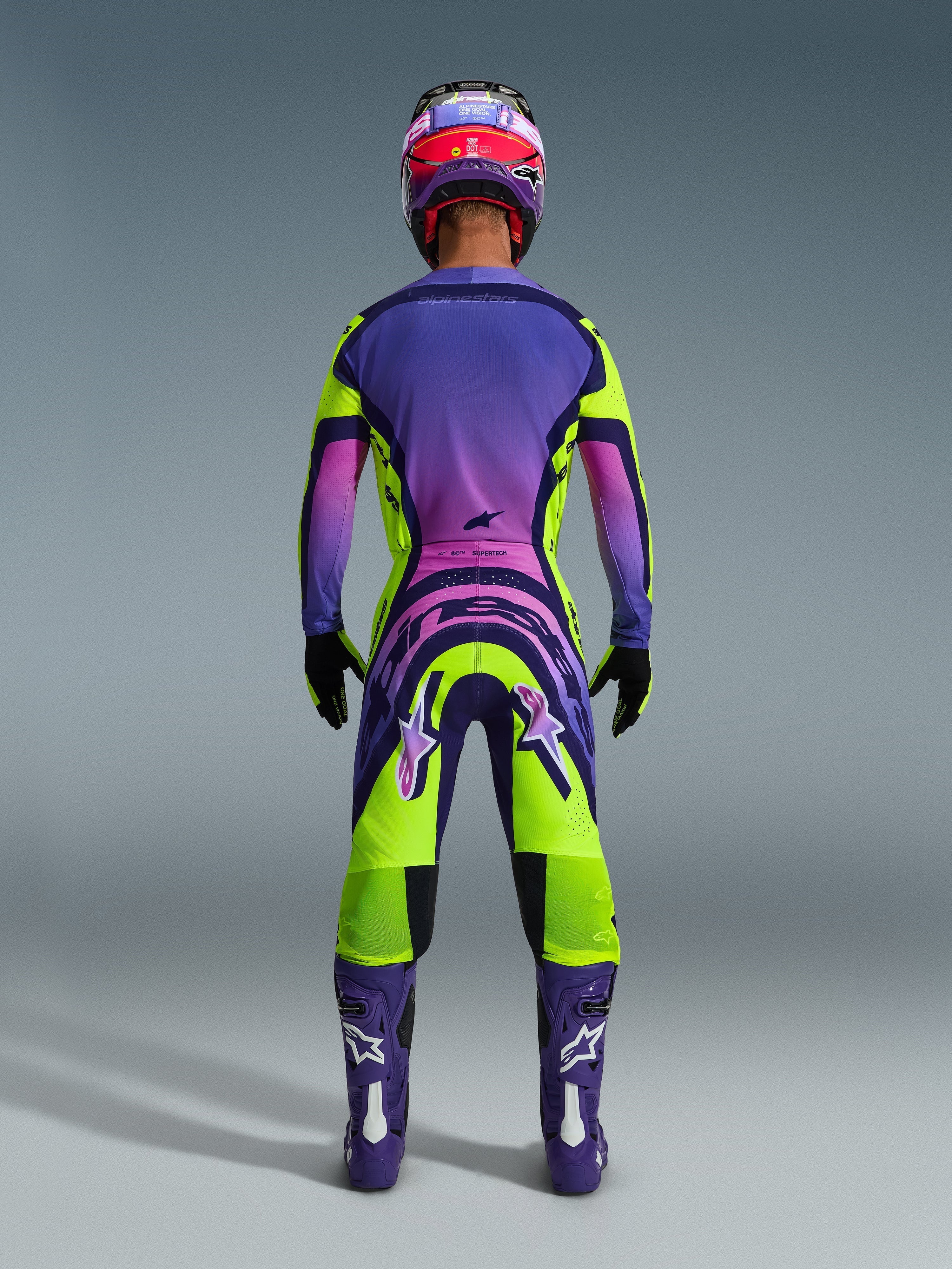Motocross rider from behind wearing the 2026 Supertech Vista Jersey and matching pants, vibrant yellow and purple design with pink gradients, purple boots with white Alpinestars logos, black gloves, and a red and purple racing helmet, professional-grade racing gear set.