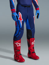 Alpinestars Pantalones Supertech Vista 2026, pantalones de motocross, azul marino oscuro, coral intenso y cobalto, usados con botas de carreras de color rojo brillante y guantes azules, con logotipos gráficos grandes en los muslos y paneles internos reforzados en negro en las rodillas para rendimiento todoterreno.