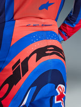 Primer plano de los pantalones Alpinestars Supertech Vista 2026, pantalones de motocross en color coral intenso y azul cobalto, con un gran gráfico negro de Alpinestars en el asiento, perforaciones de ventilación cortadas con láser y marca 'SUPERTECH' en la parte posterior de la cintura, equipamiento de competición profesional.