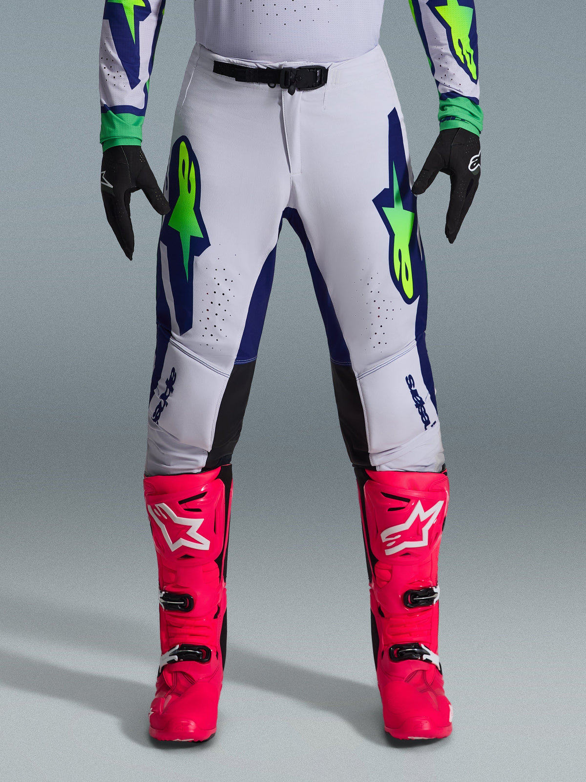 Alpinestars 2026 Supertech Vista Pants, pantalones de motocross, gris claro con detalles en amarillo flúor y azul, usados con botas rosa vibrante y guantes negros, con llamativos logotipos de estrella en los muslos y perforaciones cortadas con láser para la ventilación.