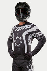 Alpinestars 2024 Racer Hana Jersey, jersey de motocross, negro y blanco, con llamativos gráficos de texto y logo de Alpinestars en blanco, usado por un piloto con casco y gafas negros, equipamiento de alto rendimiento para carreras todoterreno