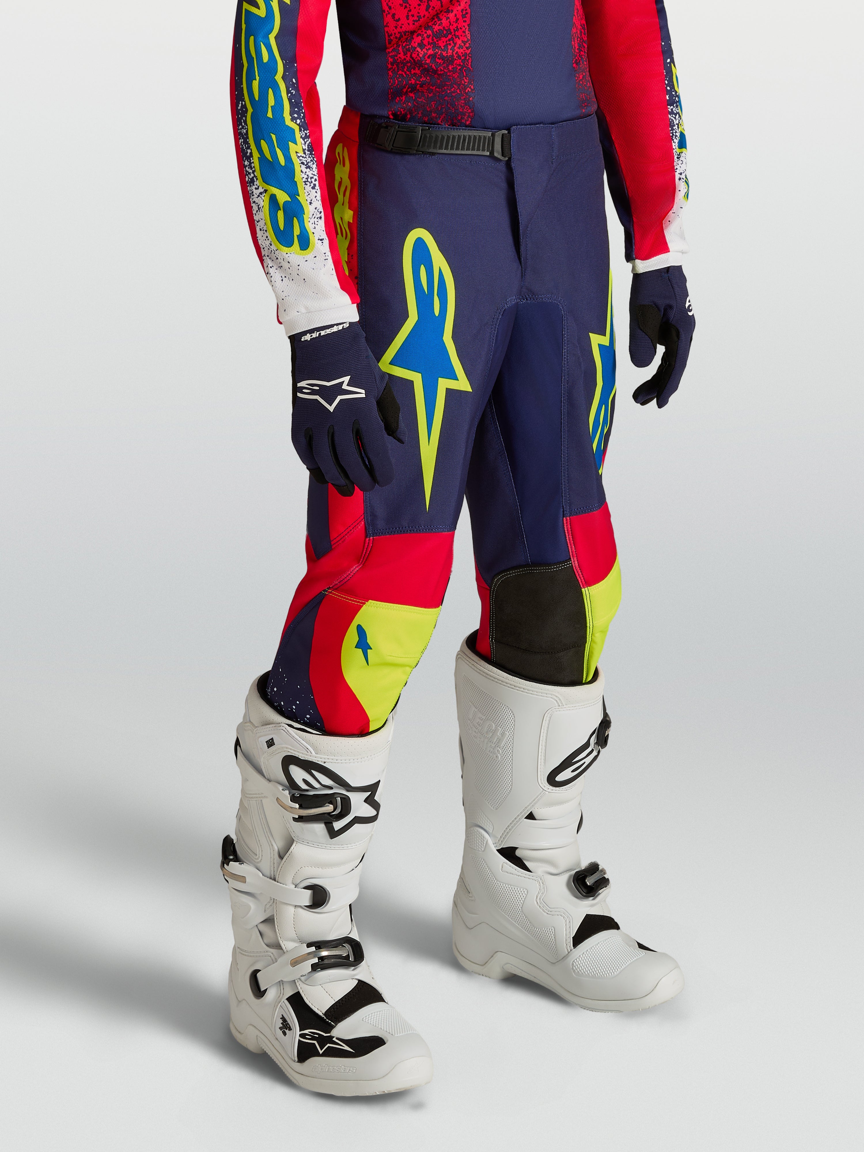 Pantalones Alpinestars Fluid Drag Juvenil de motocross, azul marino y rojo con detalles neón y botas blancas.
