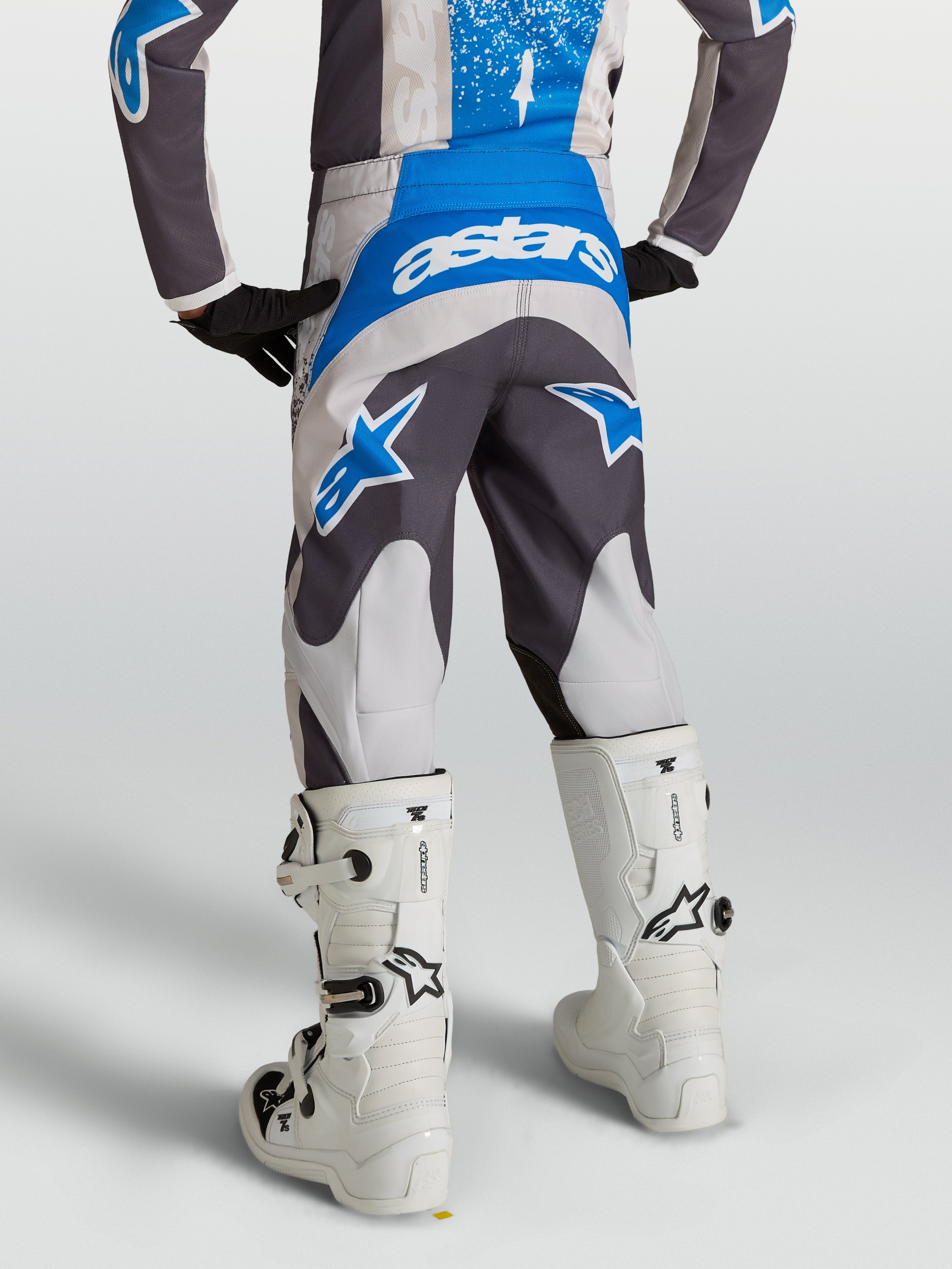 Pantalones Fluid Drag Juvenil, pantalones de motocross para niños en gris y azul, vista trasera en piloto con botas blancas, equipo técnico para conducción off-road