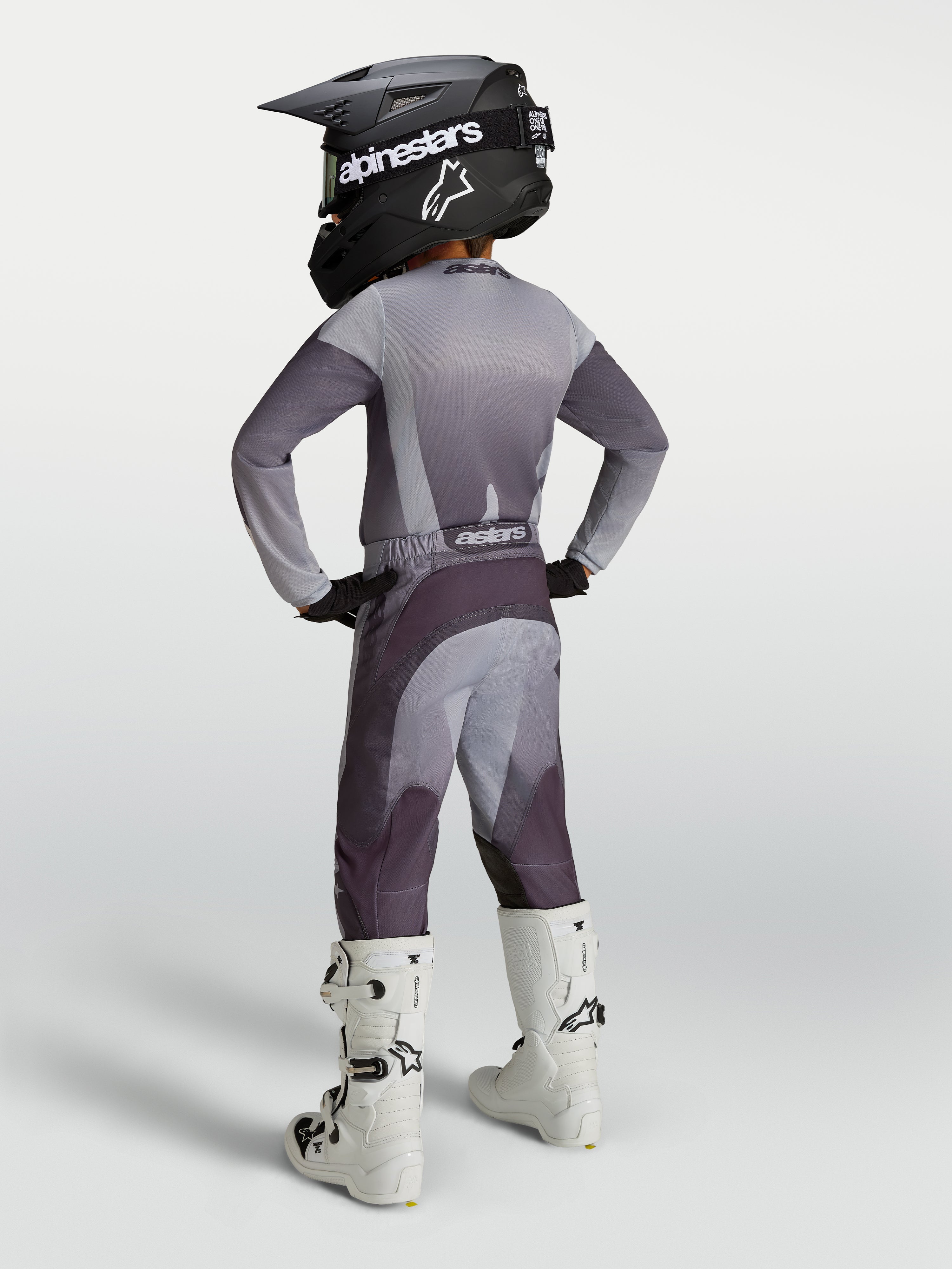 Alpinestars Pantalones Fluid Amber Juvenil, equipo de motocross gris y morado carbón, ajuste deportivo, con botas blancas.