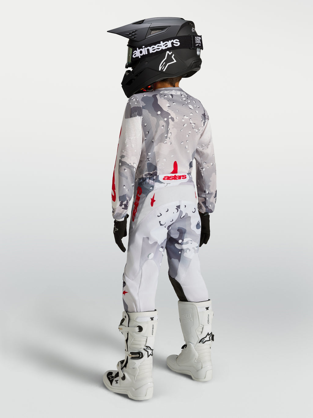 Alpinestars Pantalones Fluid Lone Juvenil, equipo de motocross gris camuflaje y blanco con detalles rojos, diseño para off-road en un piloto juvenil visto desde atrás.