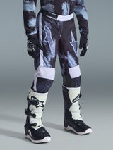 Pantalones Juveniles Alpinestars Fluid Lahnd 2026, pantalones de motocross, camuflaje Iron Camo en gris, negro y blanco, usados con jersey de manga larga a juego, guantes de carreras negros y botas de motocross blancas y negras, con grandes logotipos de estrella en los muslos.