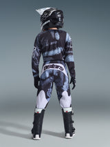 Alpinestars 2026 Youth Fluid Lahnd Pants, pantalones de motocross juveniles, Iron Camo con tonos azules y grises, vista trasera del piloto usando el conjunto de equipamiento a juego con casco y botas, acentos blancos en las piernas y logotipo 'astars' en la parte trasera.