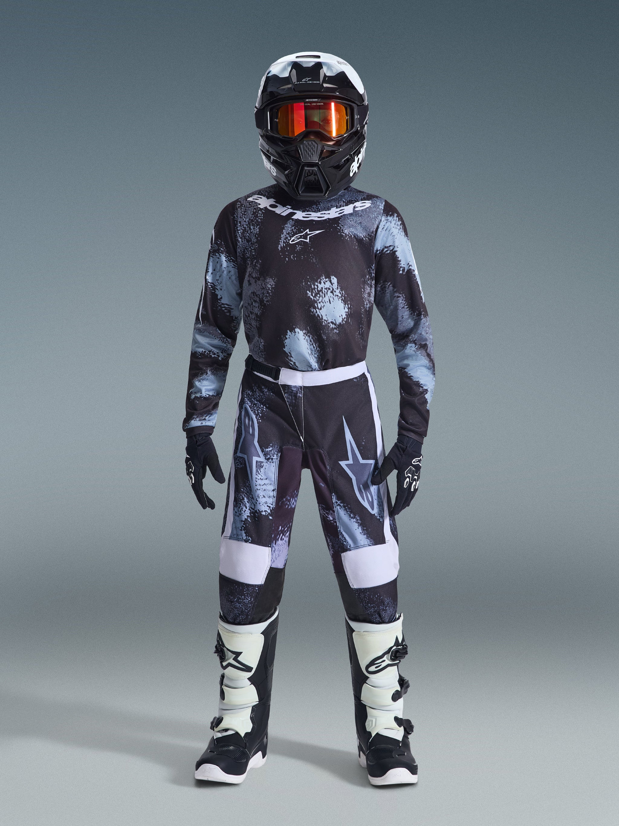 Alpinestars 2026 Youth Fluid Lahnd Pants, pantalones de motocross juveniles, Iron Camo, usados por un joven piloto con jersey de camuflaje a juego, casco negro con gafas naranjas y botas blancas y negras, con un diseño coordinado con logotipos llamativos y paneles reforzados para carreras todoterreno.
