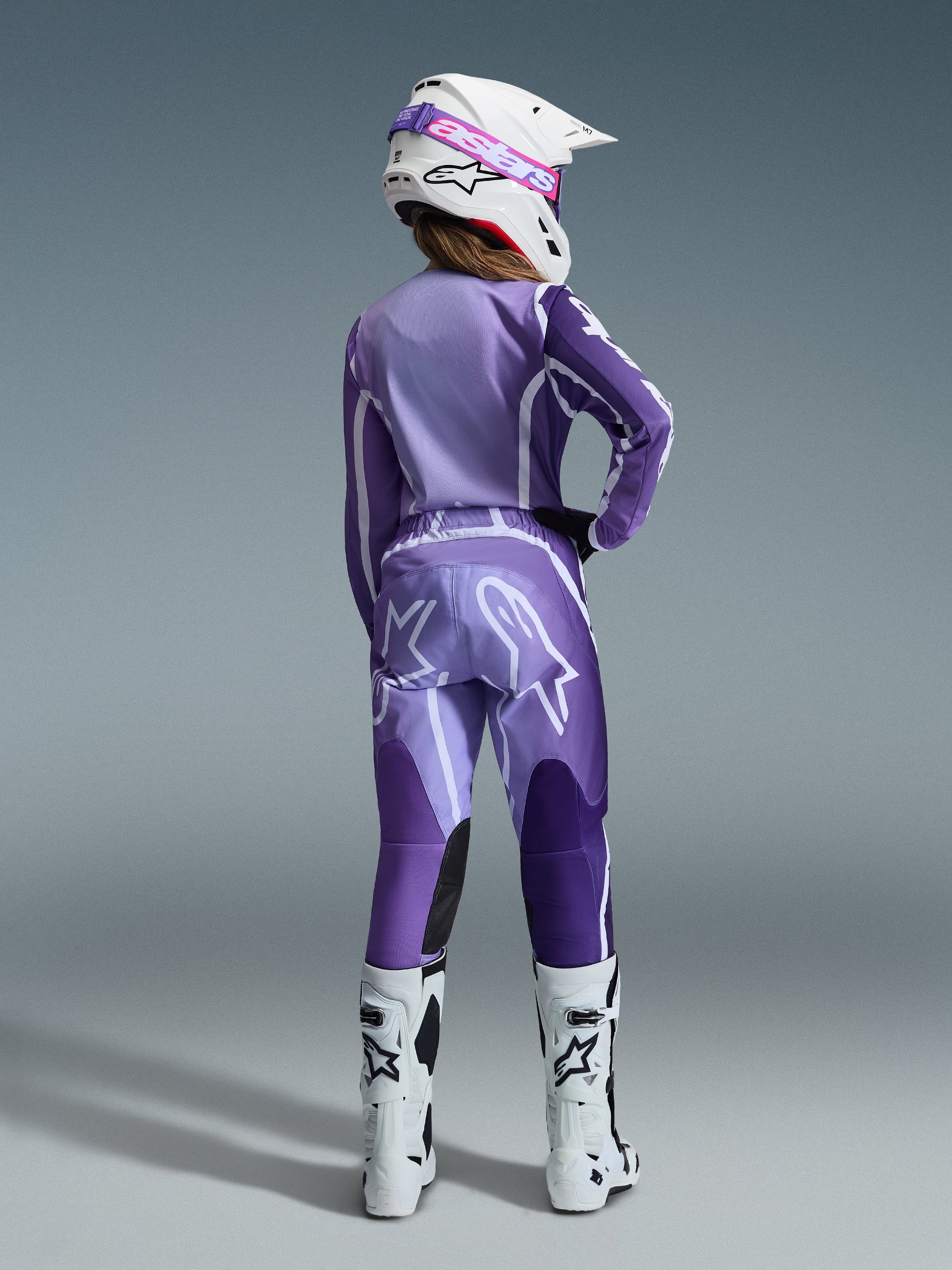 Piloto de motocross vista desde atrás con los pantalones Alpinestars Stella Fluid Apex 2026 para mujer en violeta y blanco, jersey morado de manga larga a juego, casco blanco con gafas rosas y blancas, y botas blancas de competición; conjunto de motocross coordinado.