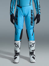 Alpinestars 2026 Stella Fluid Apex Pants para mujer, pantalones de motocross, cian brillante y negro, marca negra vertical en los muslos, paneles reforzados negros en el interior de las rodillas, mostrados con botas de motocross blancas y negras y guantes negros, equipamiento profesional de off-road para mujer