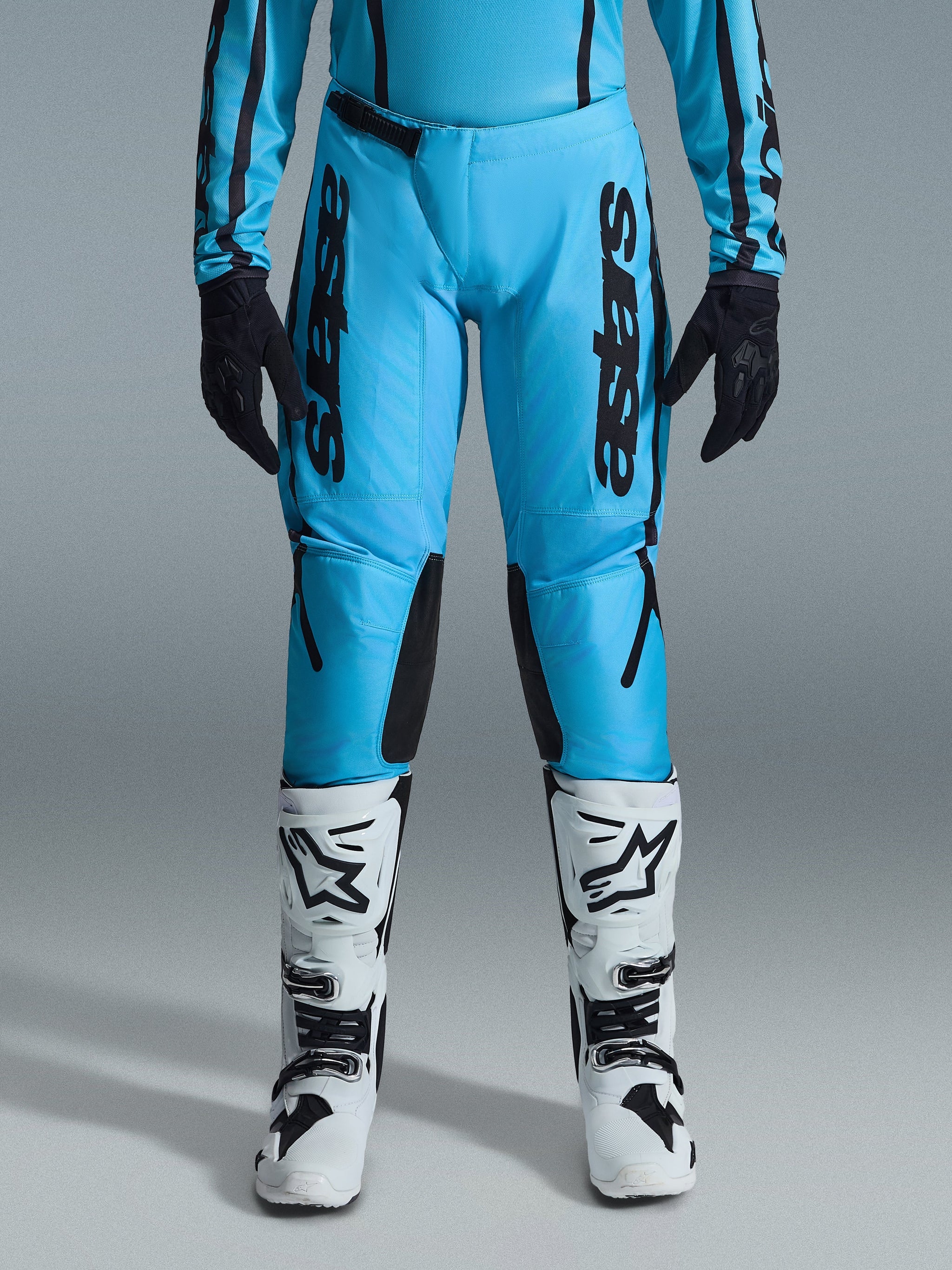 Alpinestars 2026 Stella Fluid Apex Pants para mujer, pantalones de motocross, cian brillante y negro, marca negra vertical en los muslos, paneles reforzados negros en el interior de las rodillas, mostrados con botas de motocross blancas y negras y guantes negros, equipamiento profesional de off-road para mujer