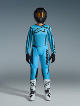Piloto de motocross con el conjunto Alpinestars Stella Fluid Apex 2026, jersey y pantalones azul cian con detalles negros, casco negro con gafas iridiscentes y botas blancas, equipamiento profesional completo de motocross