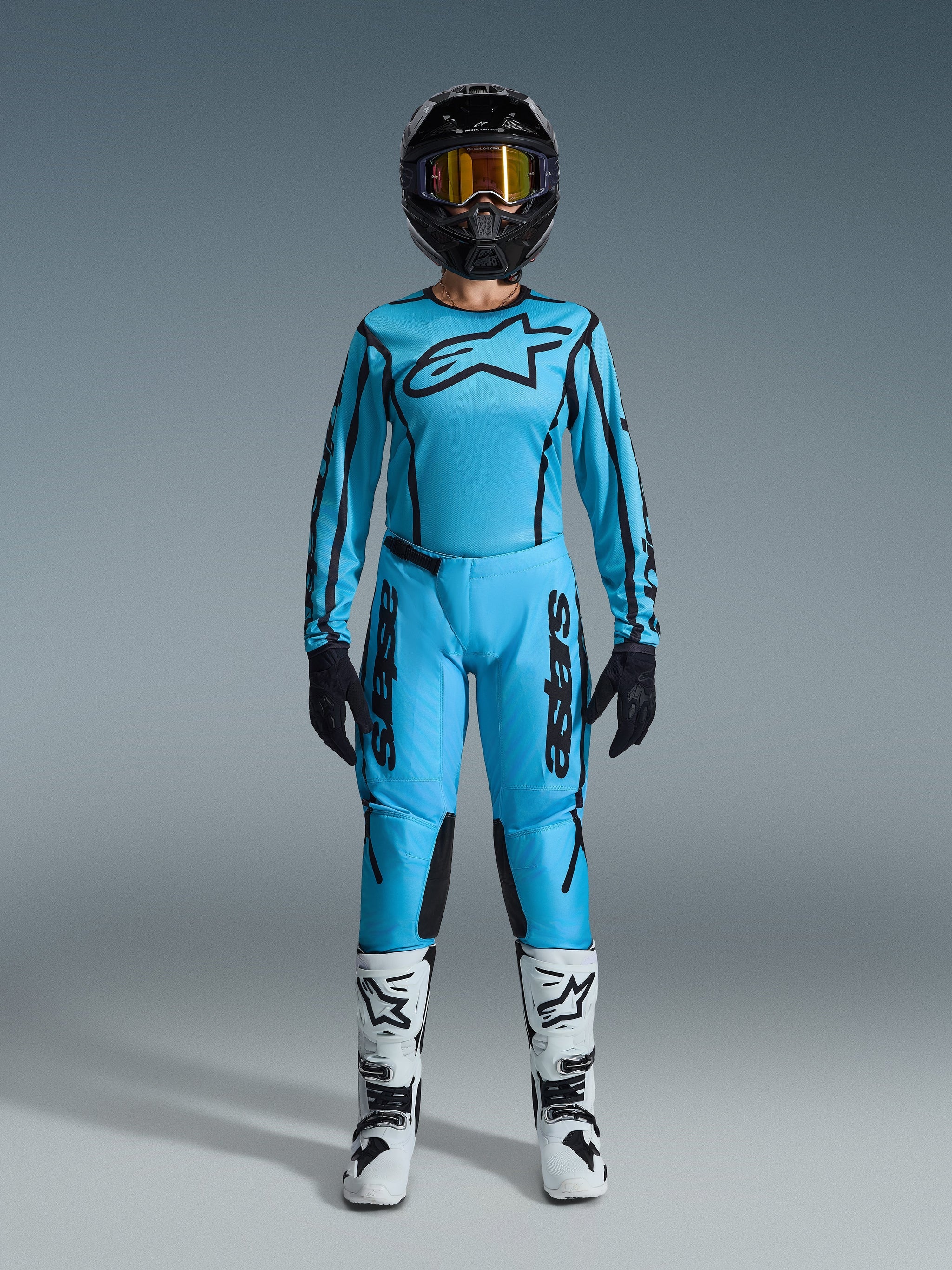 Piloto de motocross con el conjunto Alpinestars Stella Fluid Apex 2026, jersey y pantalones azul cian con detalles negros, casco negro con gafas iridiscentes y botas blancas, equipamiento profesional completo de motocross