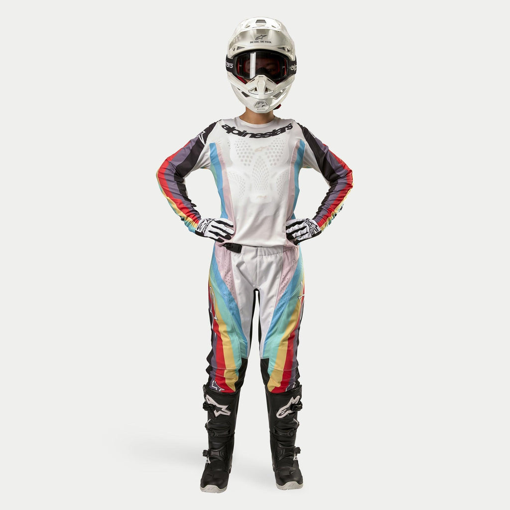 Piloto de motocross con pantalones Alpinestars Stella Techstar para mujer 2024, Negro Multicolor con base blanca y franjas rojas, amarillas y azules, jersey de manga larga a juego, casco blanco y botas negras, equipo completo de carreras para competición todoterreno