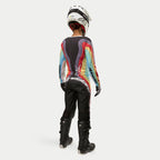 Corredora de motocross vista desde atrás vistiendo los pantalones Alpinestars Stella Techstar 2024 para mujer en Negro Multicolor, con un gran logotipo blanco en el asiento, combinados con un jersey de rayas de colores, casco blanco y botas Tech 3 negras para carreras todoterreno.