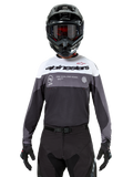 Piloto de motocross con el jersey Alpinestars Pro-Dura en Black Magnet White, camiseta técnica de manga larga con bloques de color blanco y gris, usada con casco negro y gafas, con marca 'alpinestars' y el eslogan 'ONE GOAL ONE VISION', equipamiento de competición para motociclismo off-road