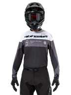 Piloto de motocross con el jersey Alpinestars Pro-Dura en Black Magnet White, camiseta técnica de manga larga con bloques de color blanco y gris, usada con casco negro y gafas, con marca 'alpinestars' y el eslogan 'ONE GOAL ONE VISION', equipamiento de competición para motociclismo off-road
