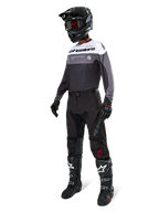 Jersey Alpinestars Pro-Dura en Black Magnet White, usado por un piloto de motocross con equipamiento completo que incluye pantalones negros, casco negro con gafas y botas blancas y negras. El jersey presenta un diseño técnico de bloques de color con paneles blancos, grises y negros, diseñado para el rendimiento en enduro y conducción off-road.