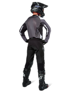 Piloto de motocross con el Jersey Alpinestars Pro-Dura en Black Magnet White, vista trasera que muestra el panel de malla gris y mangas negras con la marca Alpinestars en blanco, coordinado con pantalones negros, casco negro y botas blancas y negras, equipo profesional de enduro