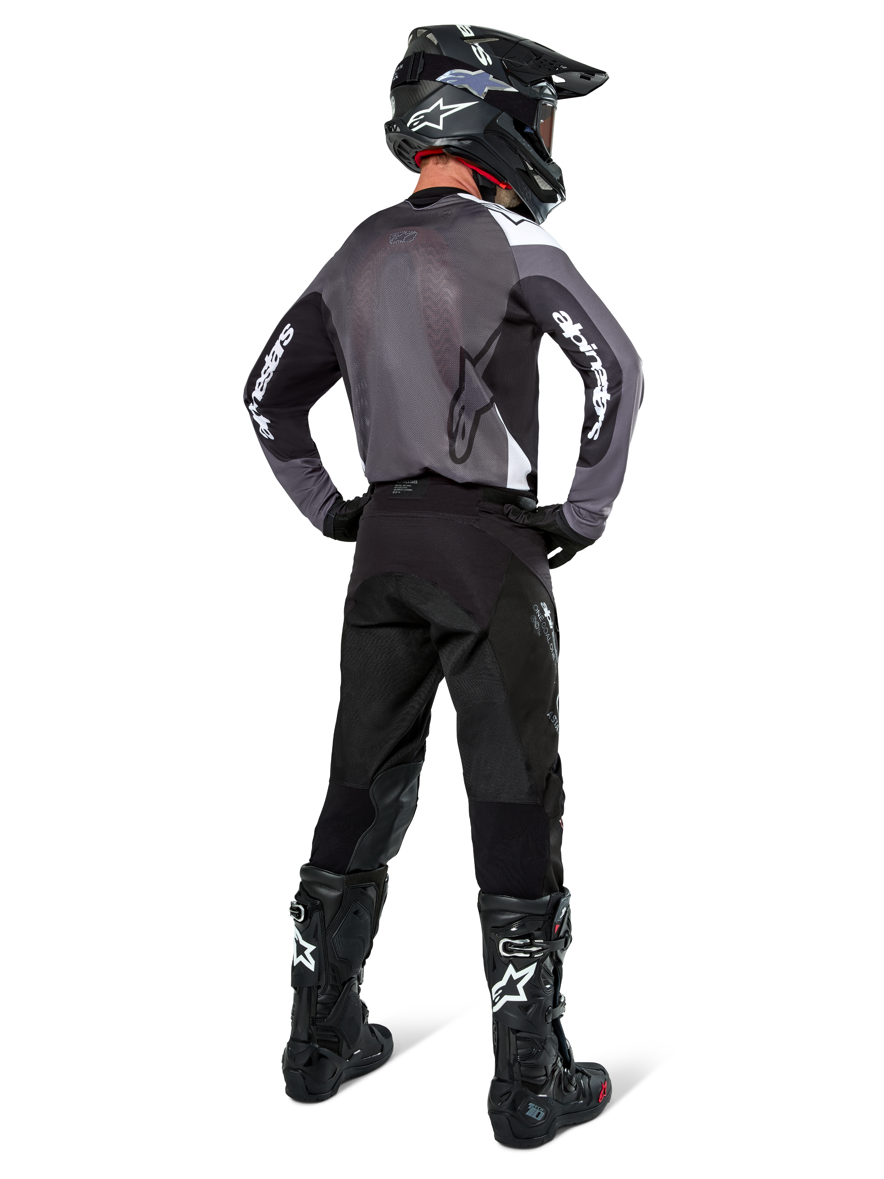 Piloto de motocross con el Jersey Alpinestars Pro-Dura en Black Magnet White, vista trasera que muestra el panel de malla gris y mangas negras con la marca Alpinestars en blanco, coordinado con pantalones negros, casco negro y botas blancas y negras, equipo profesional de enduro
