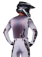 Conjunto de equipo Alpinestars Supertech Maker 2025, piloto de motocross visto desde atrás, jersey blanco y gris con logotipos negros grandes en las mangas y logo de estrella, pantalones blancos a juego con gráficos negros, casco blanco y negro, traje de carreras todoterreno profesional