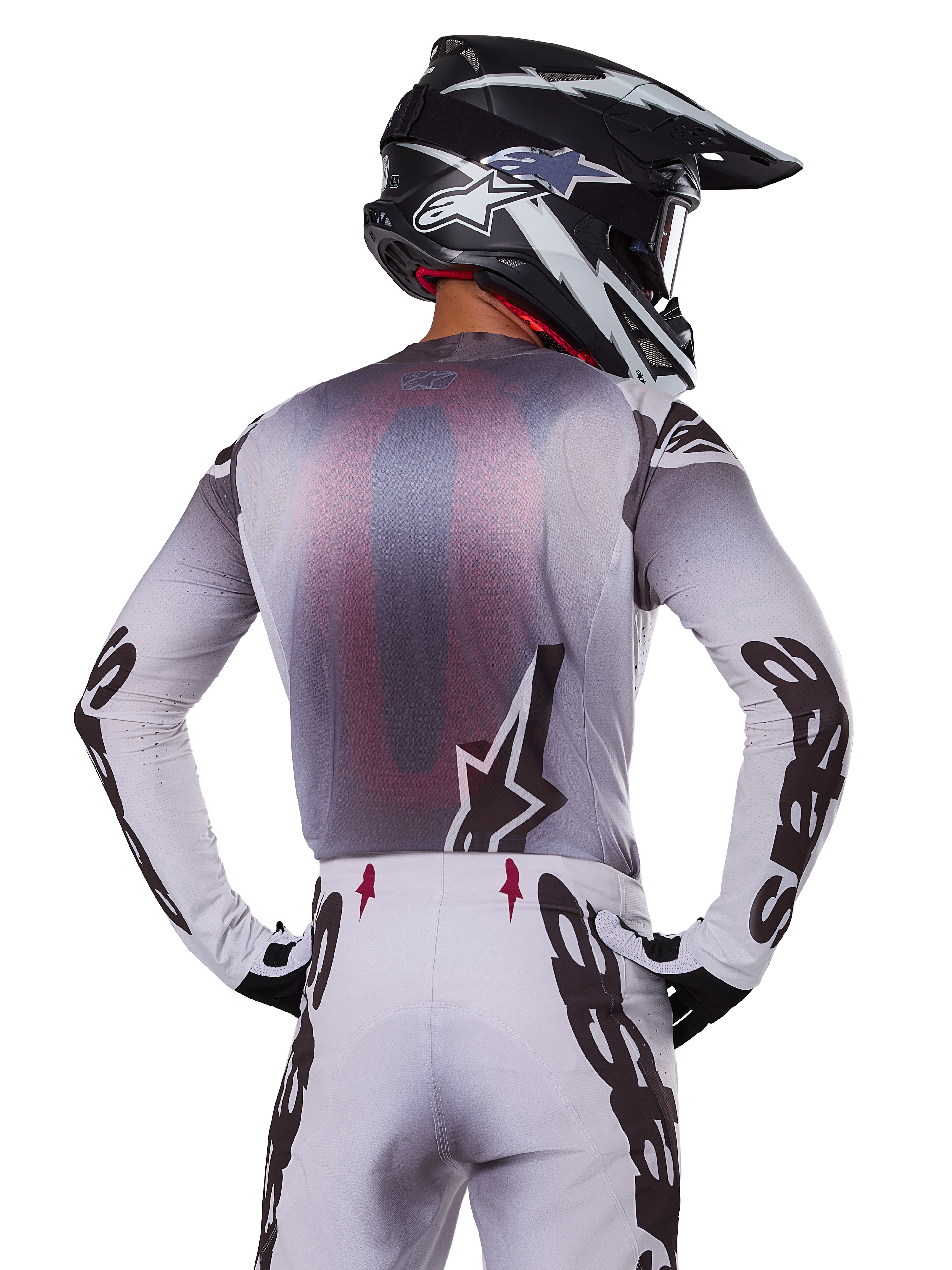 Conjunto de equipo Alpinestars Supertech Maker 2025, piloto de motocross visto desde atrás, jersey blanco y gris con logotipos negros grandes en las mangas y logo de estrella, pantalones blancos a juego con gráficos negros, casco blanco y negro, traje de carreras todoterreno profesional