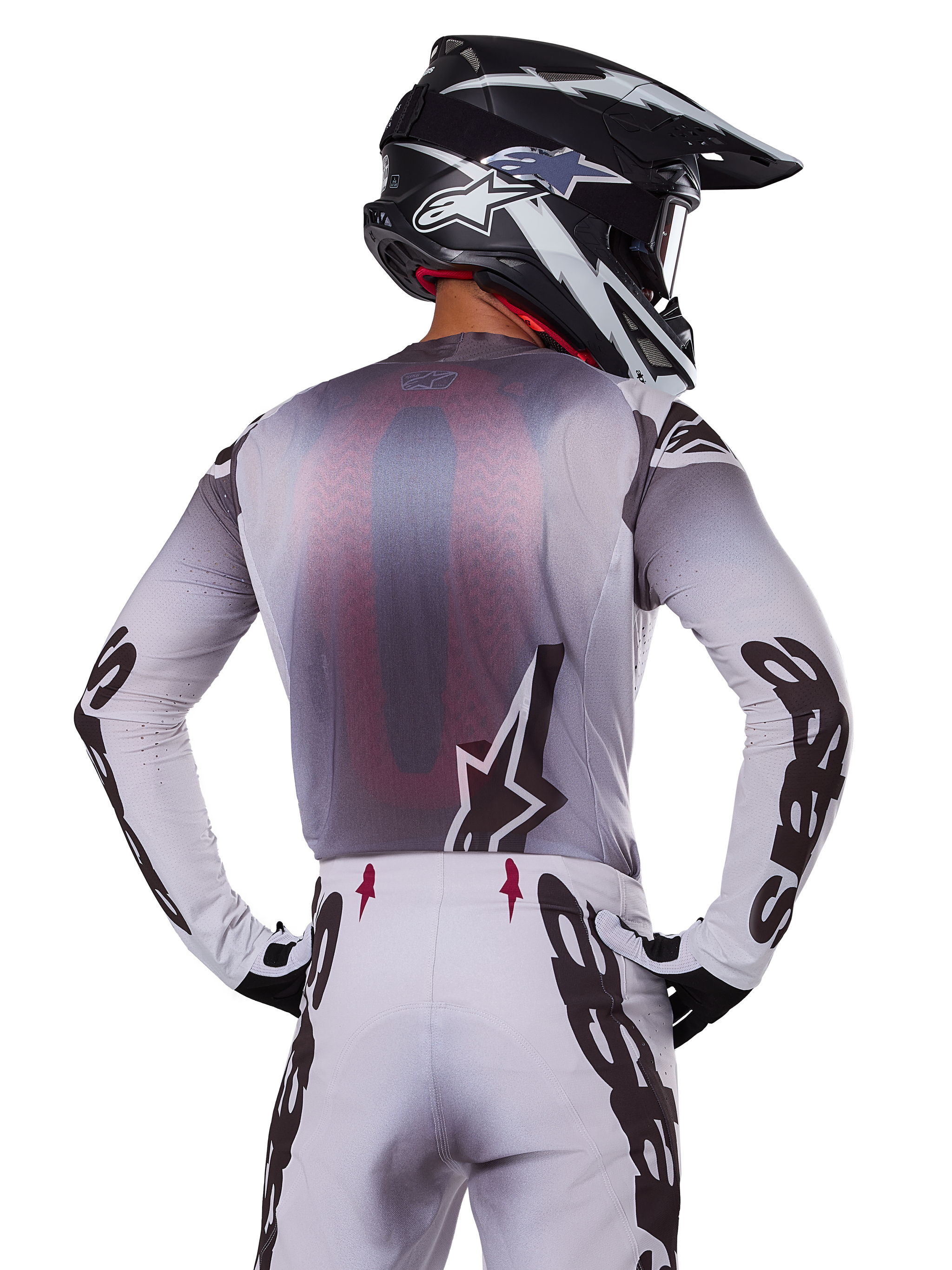 Motocross rider from behind wearing the Alpinestars 2025 Supertech Maker Jersey, Silver and Black with a red mesh back panel and black sleeve logos, paired with matching motocross pants and a black and white helmet, professional off-road racing gear