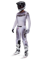 Piloto de motocross con el jersey Alpinestars 2025 Supertech Maker en plata y negro, pantalones plateados y negros a juego, casco negro y blanco con gafas negras, botas blancas y negras, y guantes coordinados, con un diseño de degradado profesional para carreras todoterreno