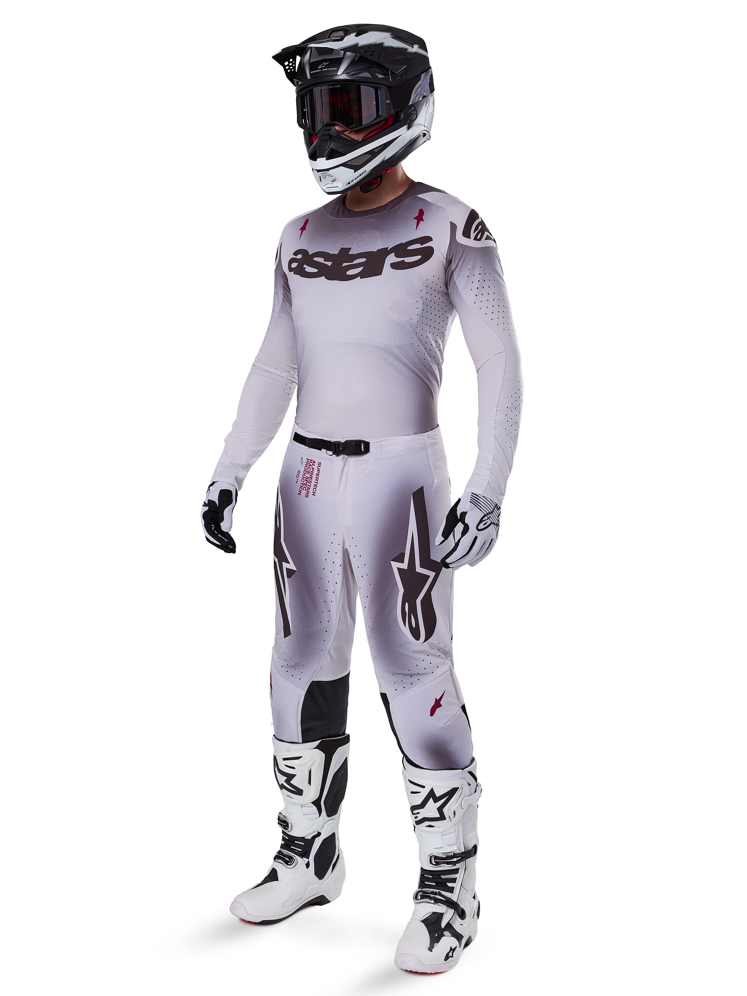 Piloto de motocross con el jersey Alpinestars 2025 Supertech Maker en plata y negro, pantalones plateados y negros a juego, casco negro y blanco con gafas negras, botas blancas y negras, y guantes coordinados, con un diseño de degradado profesional para carreras todoterreno