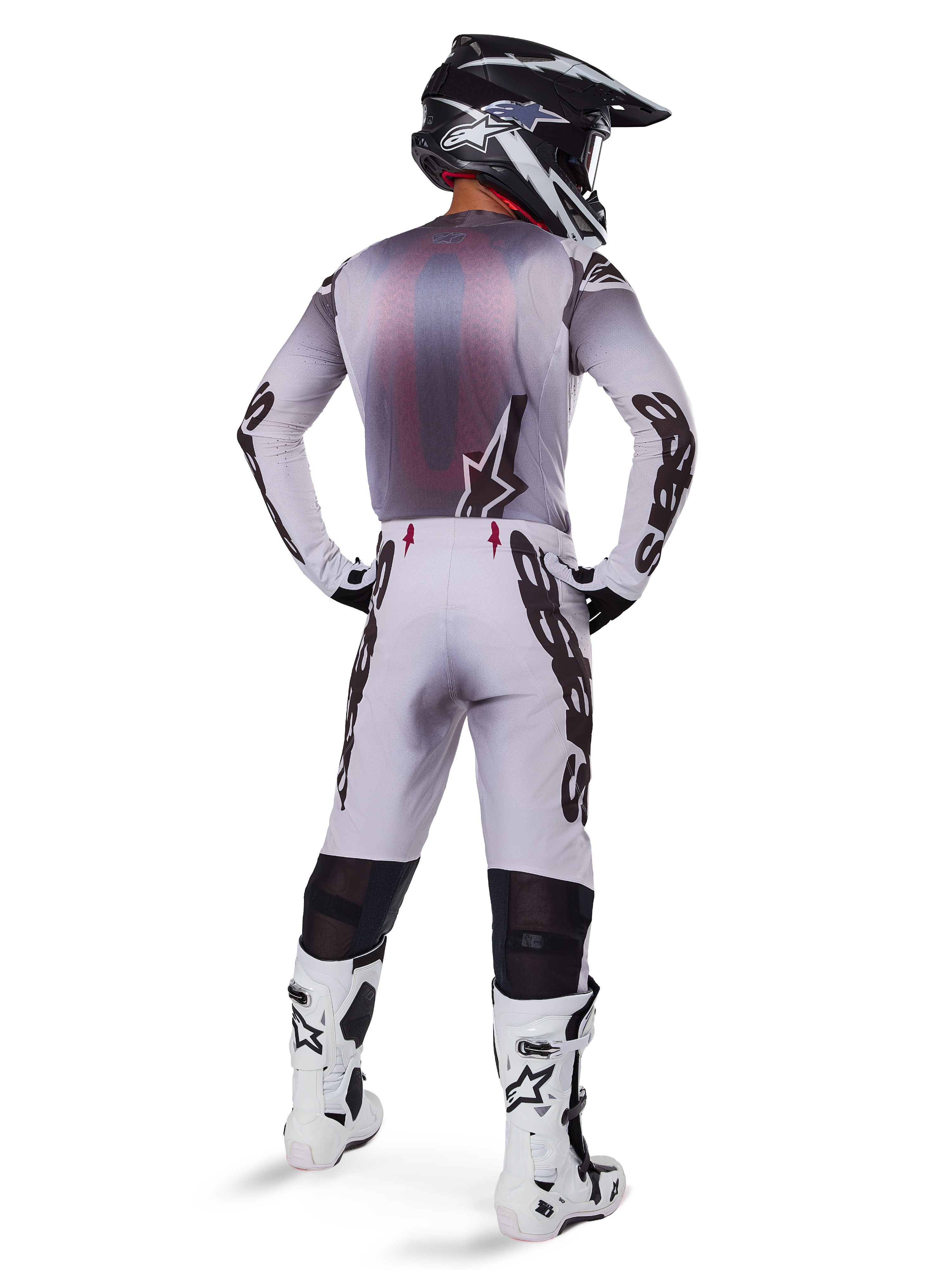 Piloto de motocross visto desde atrás con pantalones Alpinestars 2025 Supertech Maker en plata y negro, jersey de manga larga a juego, casco negro y blanco, y botas blancas y negras, con logotipos negros llamativos en las piernas, equipamiento profesional de carreras off-road