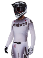 Piloto de motocross con el conjunto de equipamiento Alpinestars 2025 Supertech Maker en blanco, con jersey con degradado gris, logotipo astars en negro y perforaciones cortadas con láser, pantalones a juego con hebilla magnética y gráficos de estrellas, usado con casco y guantes en blanco y negro para un look completo de competición off-road.