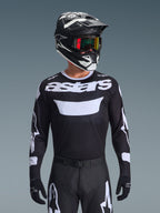Alpinestars 2026 Racer Air Riway Jersey, jersey de motocross en blanco y negro con marca 'astars', usado por un piloto con casco, gafas y pantalones negros a juego, con paneles de malla transpirable para carreras todoterreno