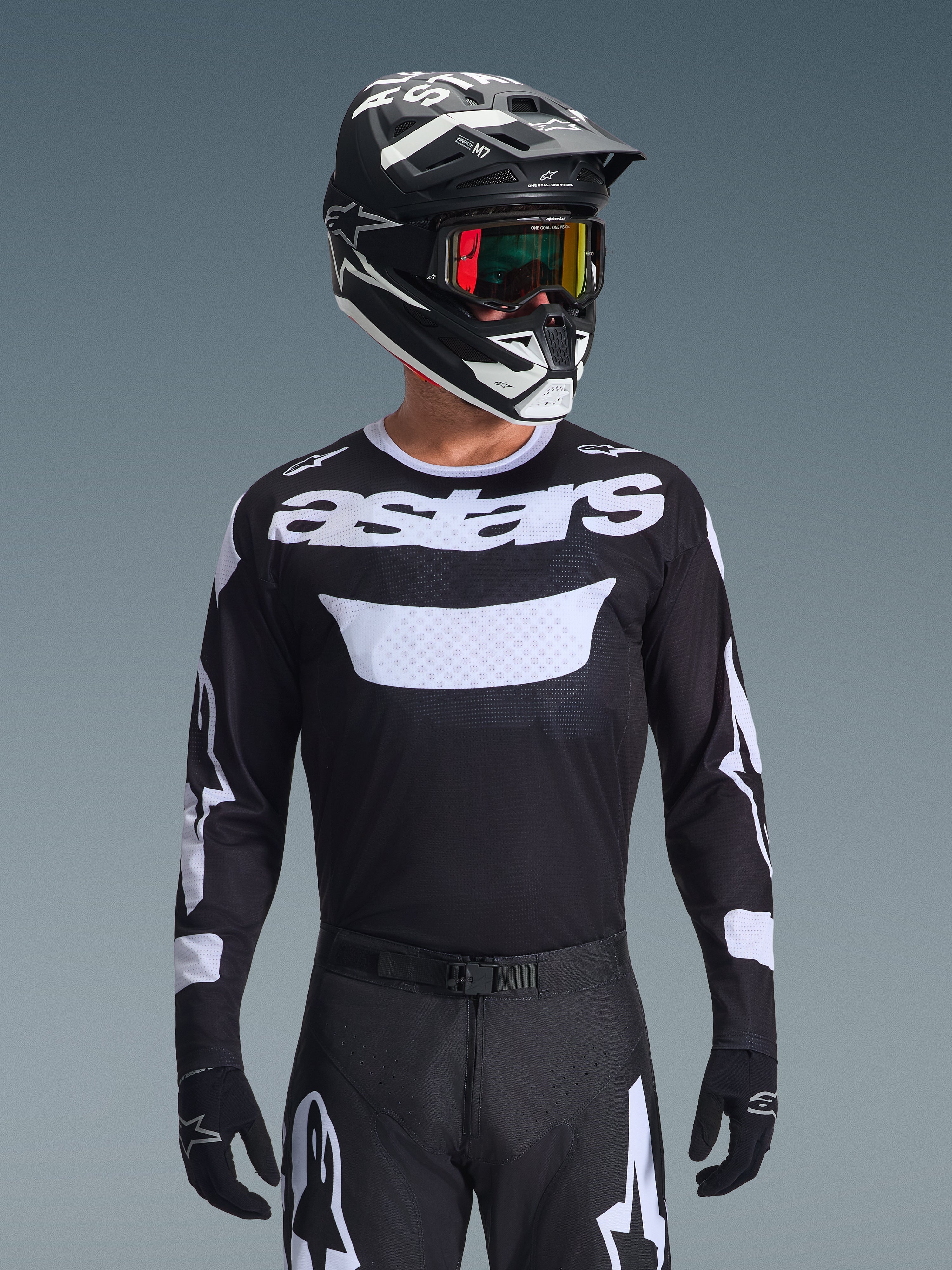 Piloto de motocross con el conjunto de equipo Alpinestars 2026 Racer Air Riway, jersey negro con logo 'astars' blanco y panel ventilado en el pecho, pantalones negros con detalles blancos, casco a juego en blanco y negro con gafas iridiscentes, guantes negros, equipo de motocross de alto rendimiento en blanco y negro.