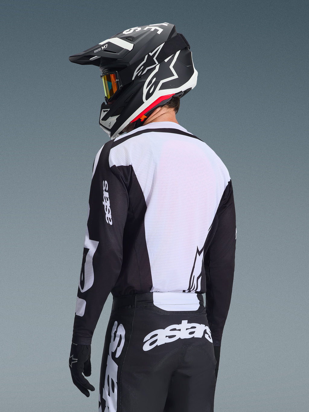 Piloto de motocross desde atrás vistiendo el Jersey Alpinestars Racer Air Riway 2026, negro y blanco, con un panel trasero de malla blanca transpirable y mangas negras con la marca 'astars', pantalones negros a juego con logotipos blancos, casco negro con detalles blancos, equipamiento profesional de off-road para un alto rendimiento en temperaturas elevadas.