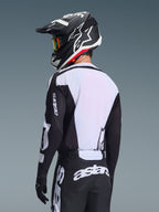Piloto de motocross desde atrás vistiendo el Jersey Alpinestars Racer Air Riway 2026, negro y blanco, con un panel trasero de malla blanca transpirable y mangas negras con la marca 'astars', pantalones negros a juego con logotipos blancos, casco negro con detalles blancos, equipamiento profesional de off-road para un alto rendimiento en temperaturas elevadas.