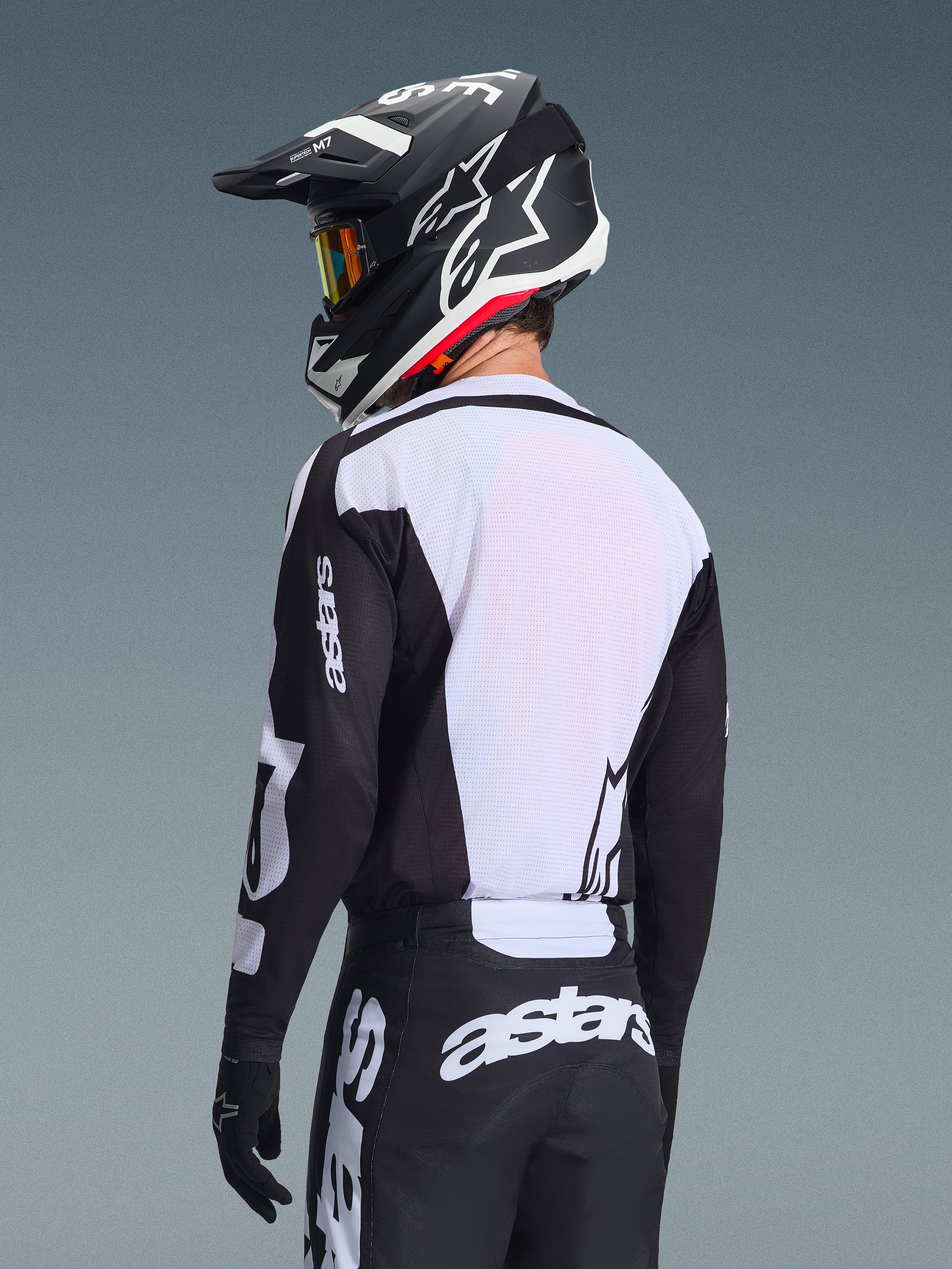 Piloto de motocross desde atrás vistiendo el Jersey Alpinestars Racer Air Riway 2026, negro y blanco, con un panel trasero de malla blanca transpirable y mangas negras con la marca 'astars', pantalones negros a juego con logotipos blancos, casco negro con detalles blancos, equipamiento profesional de off-road para un alto rendimiento en temperaturas elevadas.