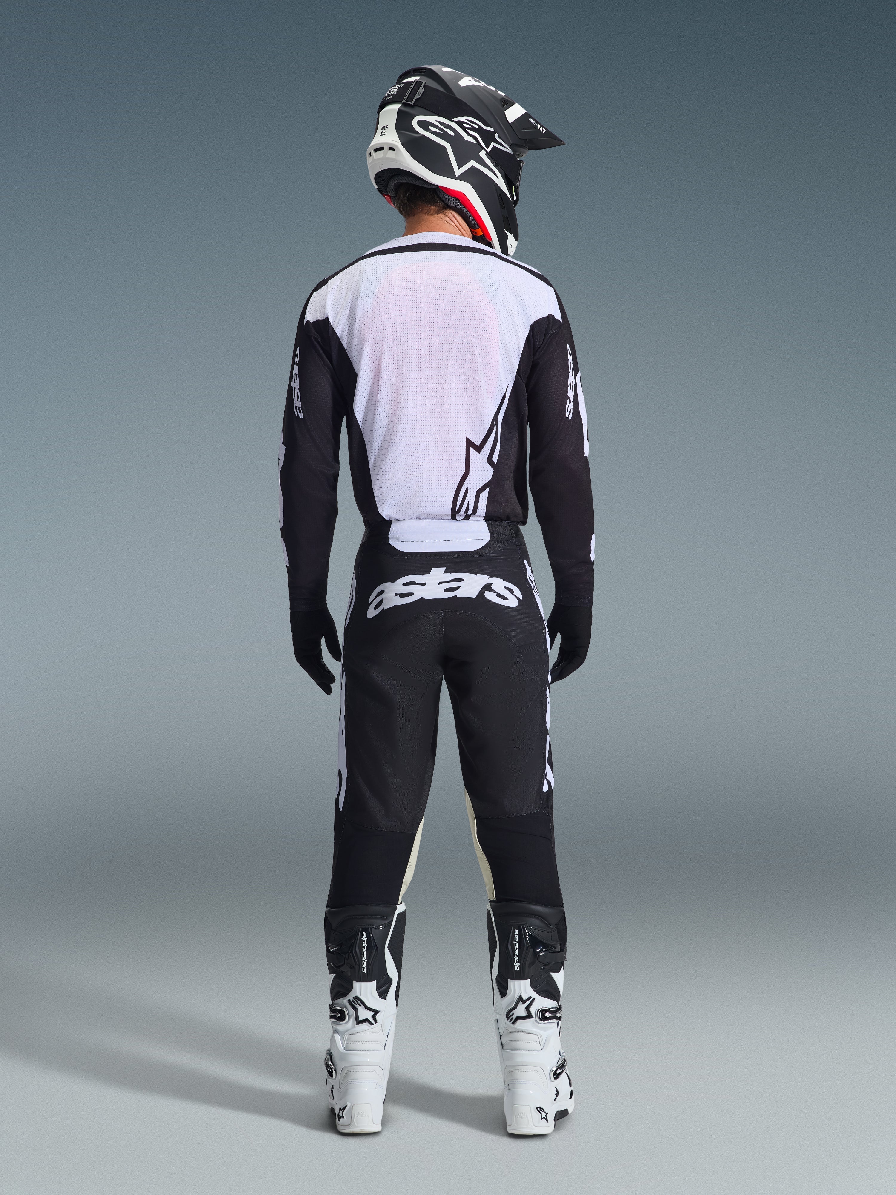 Vista trasera de un piloto de motocross con el Jersey Racer Air Riway 2026 en negro y blanco, que cuenta con un panel trasero de malla transpirable, combinado con pantalones de motocross negros con la marca 'astars' en blanco, casco negro y blanco y botas de carreras blancas.